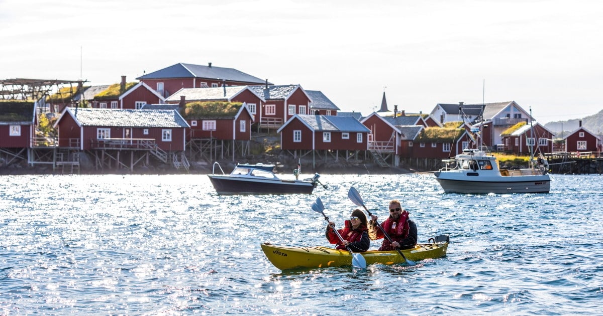 To personer padler i en gul kajakk på sjøen foran tradisjonelle røde rorbuer og båter ved kai i et nordnorsk kystsamfunn.