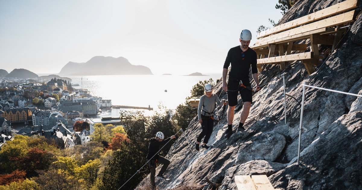 En gruppe personer med hjelm og klatreutstyr beveger seg langs en via ferrata-rute i fjellsiden over Ålesund, med byen, havet og øyene i bakgrunnen.