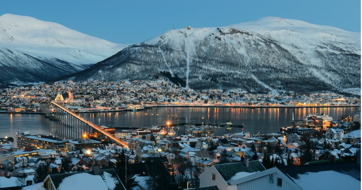 En by med mange hus ved vannet om kvelden, med snødekket fjell i bakgrunnen. Foto.