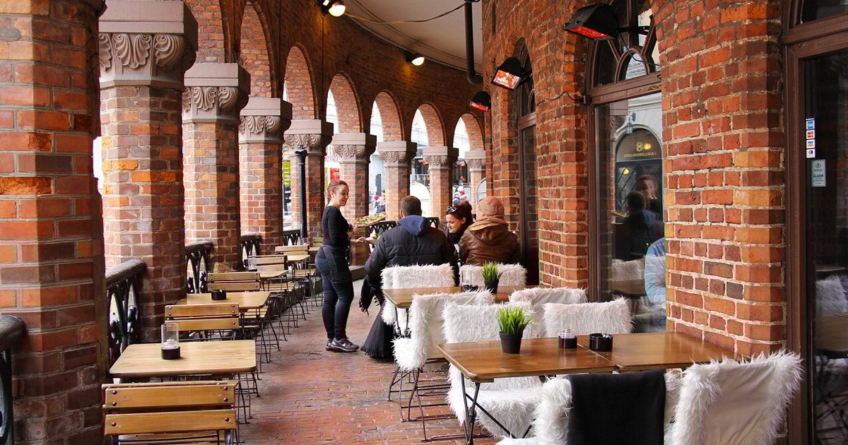 En servitrise som serverer mat til gjestene som sitter på et bord på uteservering. Foto.