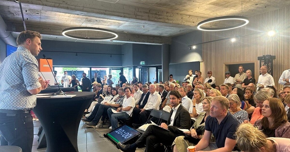 En mann står på en liten scene og holder innlegg foran en fullsatt sal. Publikum følger med, og det er både sittende og stående tilskuere. Lokalet har moderne taklamper og et minimalistisk interiør