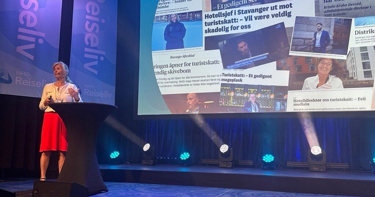Kristin Krohn Devold står på scenen under NHO Reiselivs årskonferanse 2025 og holder innlegg foran et stort lerret med avisutklipp om turistskatt. Hun står ved et høyt bord med NHO Reiseliv-logoen i bakgrunnen.