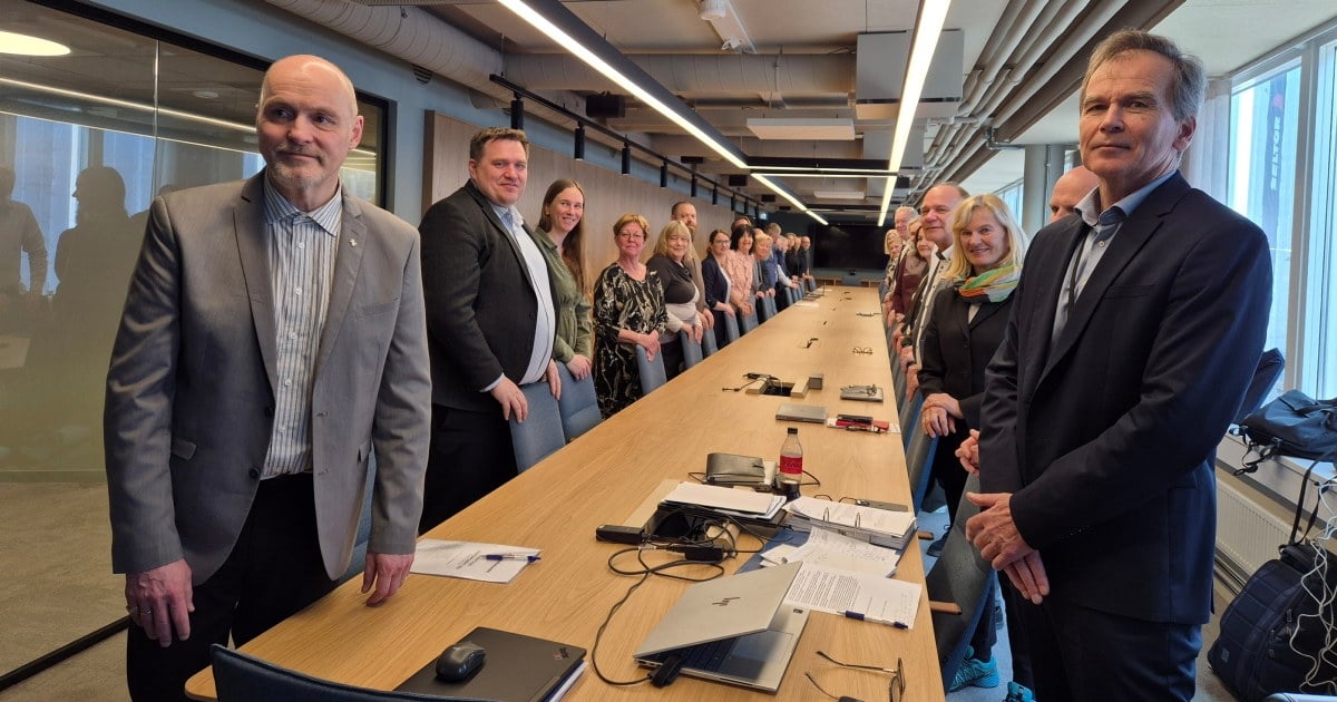 En gruppe kvinner og menn står langs begge sider av et langt møtebord i et moderne møterom. På bordet ligger laptop, dokumenter, mobiltelefoner og notatblokker. Store vinduer slipper inn dagslys fra høyre side av rommet.