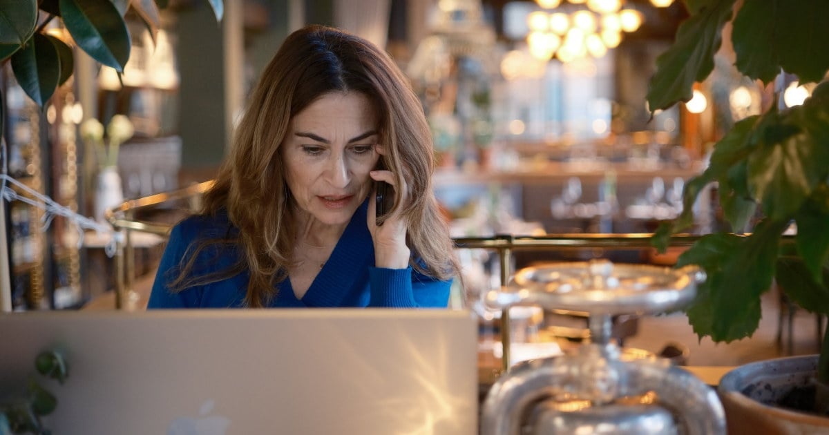 Person står ved disk i restaurantlokale og snakker i mobiltelefon mens hun arbeider på bærbar datamaskin.