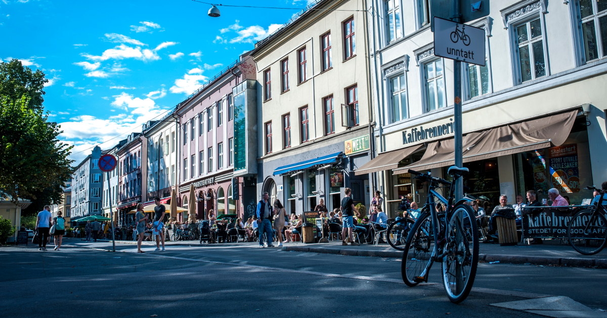 Gatebilde fra Grünerløkka i Oslo med uteservering og mennesker som nyter mat og drikke i sola. Fortauskafeer og små butikker ligger tett i tett langs bygårdene, og sykler er parkert langs fortauskanten. En blå himmel med noen skyer i bakgrunnen.