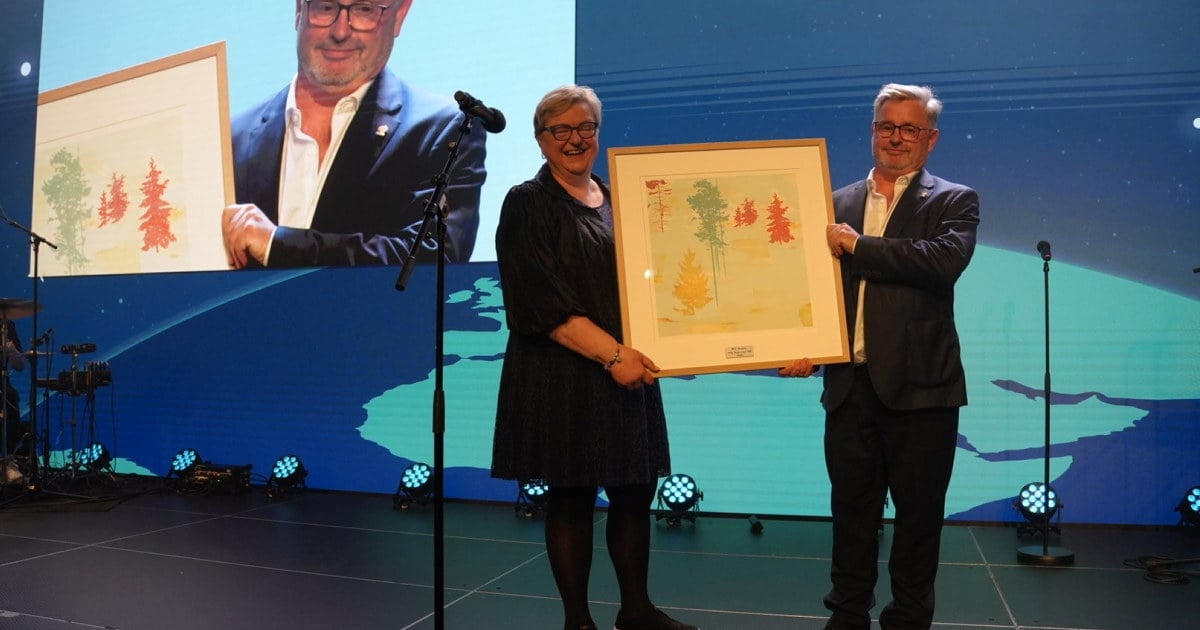 To personer står på en scene og holder et innrammet bilde sammen. I bakgrunnen vises et stort skjermbilde av personen til høyre, samt en blå scenebakgrunn med lys, mikrofonstativer og sceneteknikk.