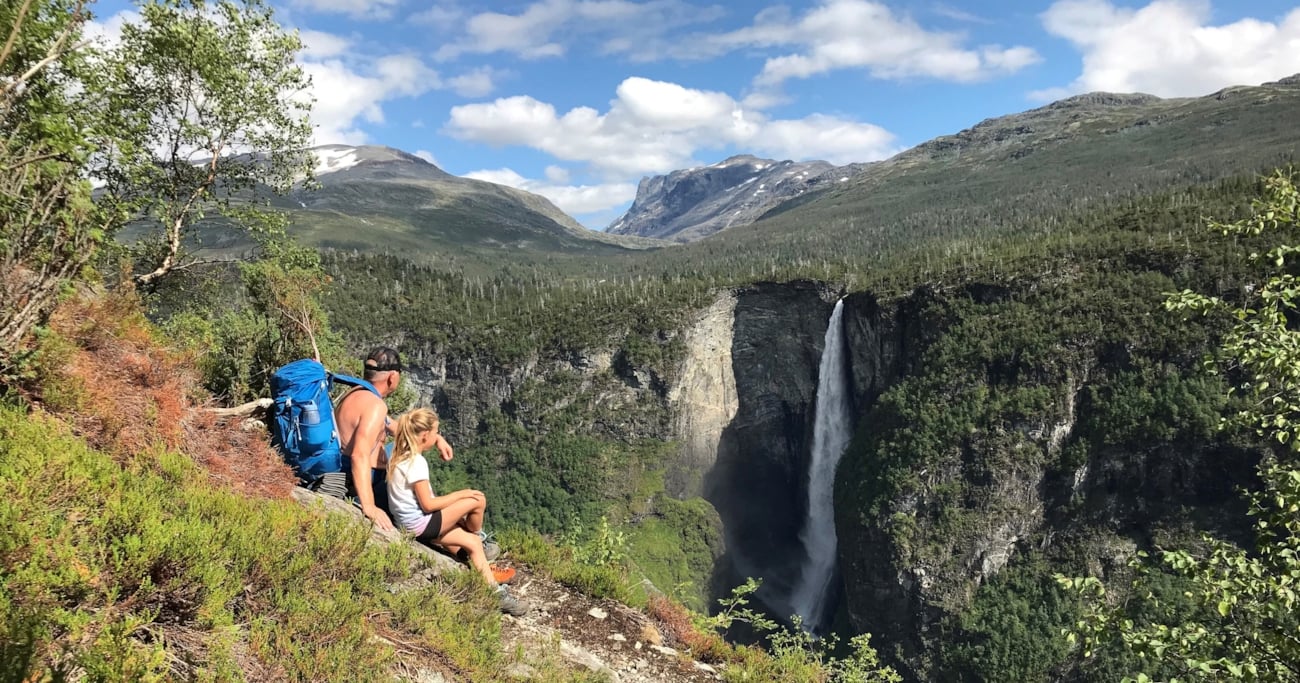 En mann i bar overkropp med ryggsekk, og en ung jente som sitter i et fjellandskap og ser mot en foss. Foto