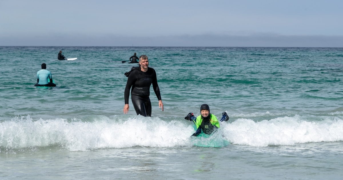Flere personer i våtdrakt surfer og bader i bølgene ved en strand. Foto.