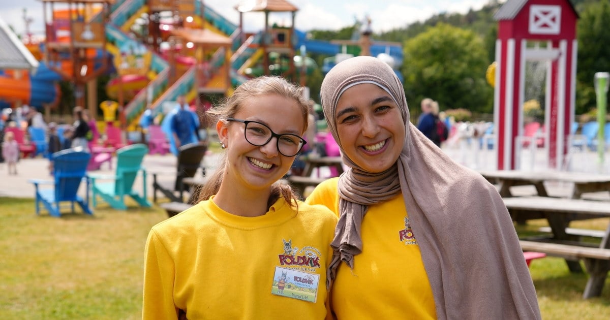 To personer i gule Foldvik Familiepark-gensere står tett sammen og smiler, med et fargerikt vannlekeområde i bakgrunnen.