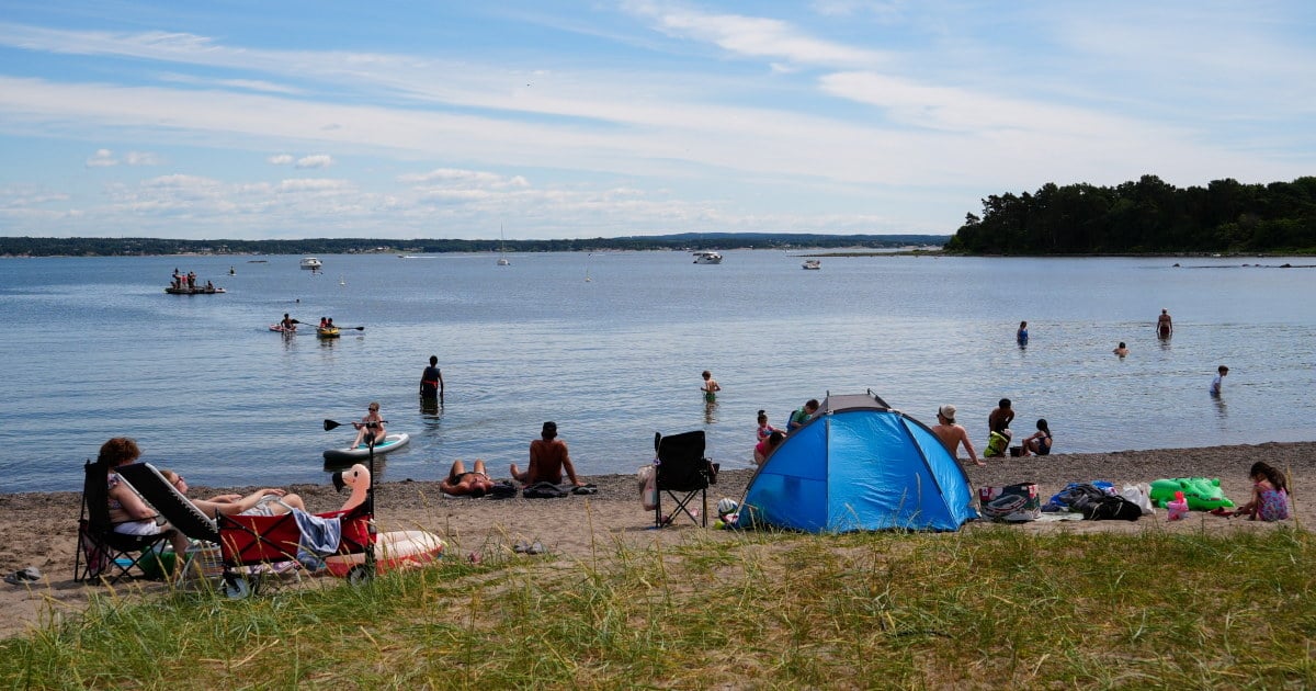 Flere personer oppholder seg på stranden og i vannet. Et blått strandtelt står på sanden, og båter sees lenger ute.