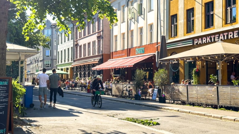 Folk sitter på uteserveringer i en solfylt bygate med fargerike bygårder, trær og forbipasserende.