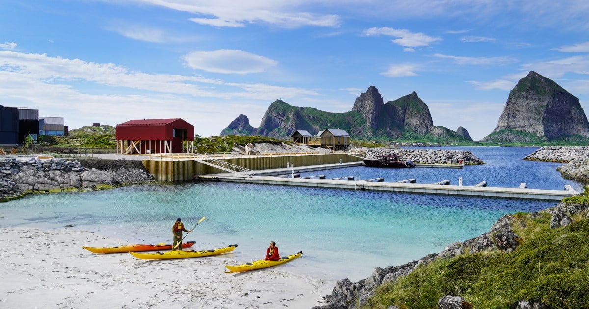 To kajakkpadlere på en lys strand, med turkist vann, småbåthavn og bratte fjell i bakgrunnen.