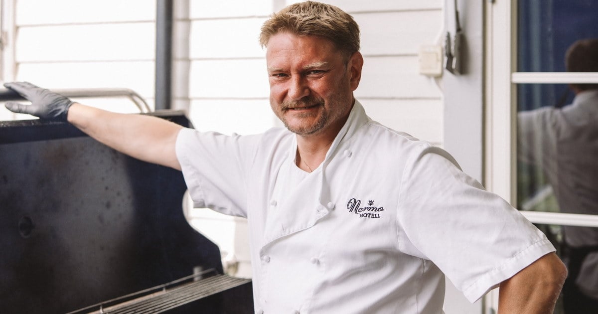 Kokk i hvit kokkejakke og svarte hansker står ved en stor grill med kjøttstykker, utenfor en hvitmalt bygning.