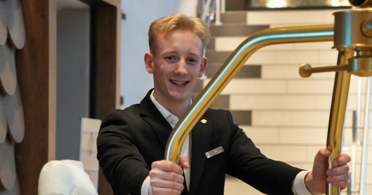 Smilende hotellansatt i uniform holder i en gyllen bagasjetralle med en opplyst trapp i bakgrunnen.