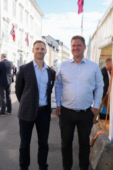 To personer står sammen og smiler i en gate, med folkeliv og arrangementstelt i bakgrunnen.