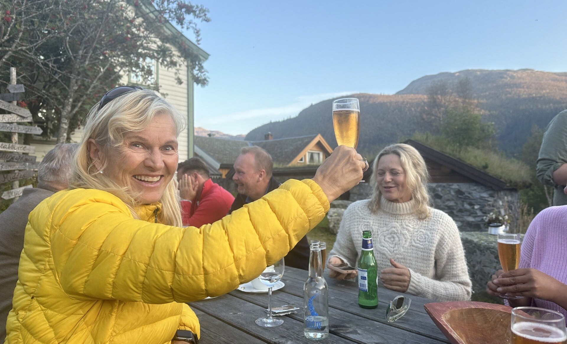 En kvinne i gul jakke skåler med et glass utendørs sammen med en gruppe mennesker, med fjell og trehus i bakgrunnen. Foto.