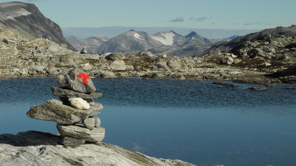 Varde med rød T-merking ved et fjellvann, omgitt av steinete fjellandskap. Spisse fjell i bakgrunnen. Foto.