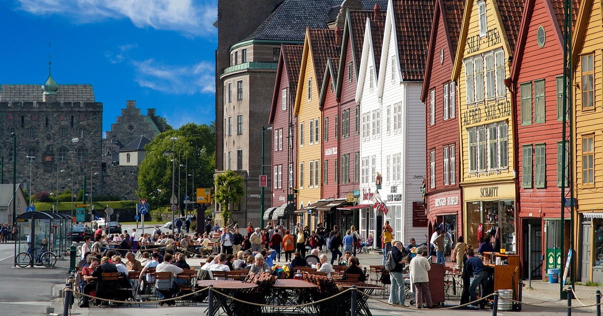 Fargerike trehus med spisse tak langs en gate, med uteservering og mange mennesker i forgrunnen.