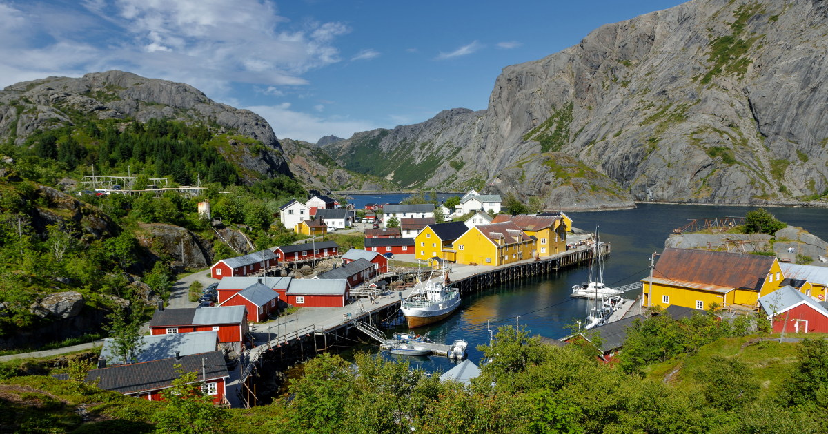 Røde og gule bygninger ved havnen i Nusfjord, med båter, sjø og fjell rundt.