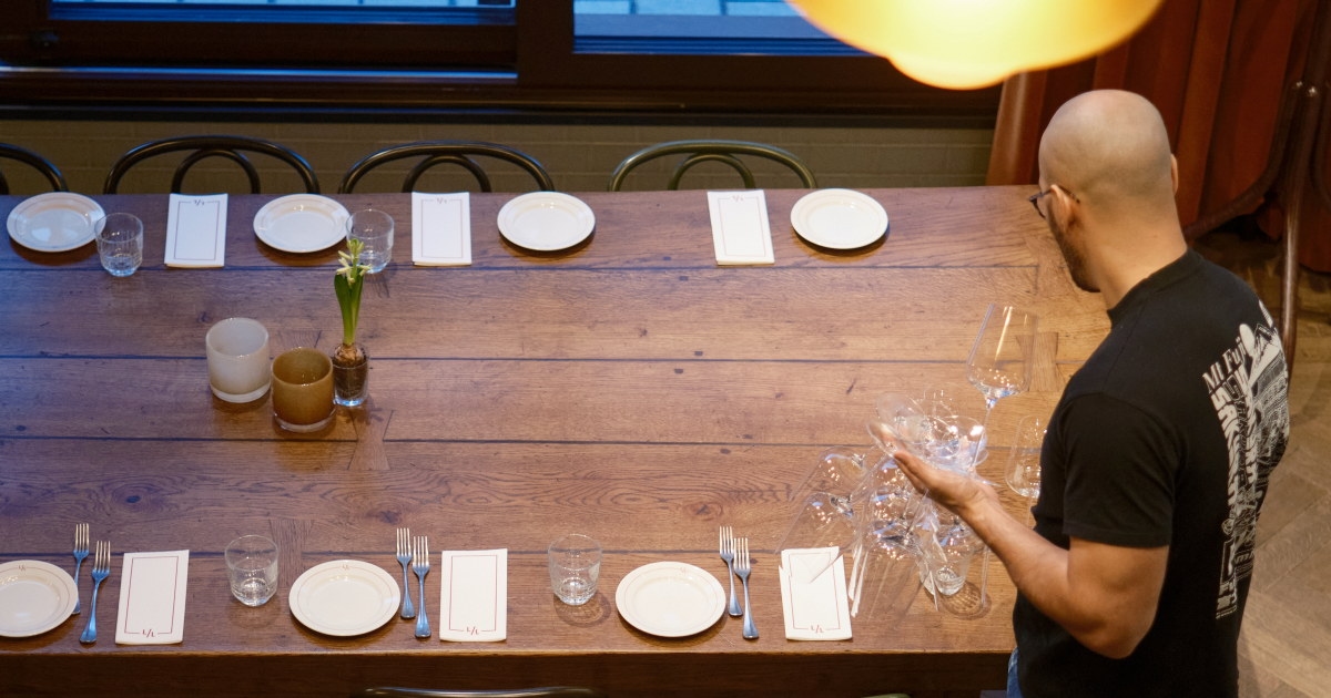 Person dekker langbord med glass og servise i restaurantlokale.