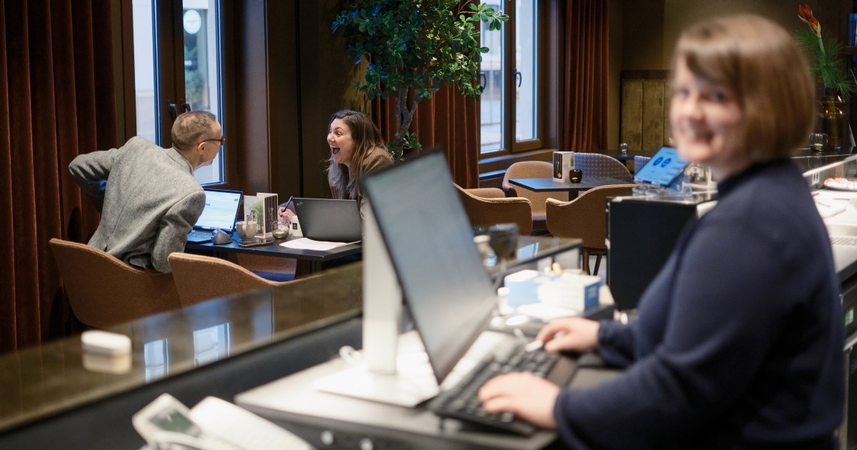 Person står i resepsjonen og arbeider ved en datamaskin, mens to personer sitter ved et bord i bakgrunnen.