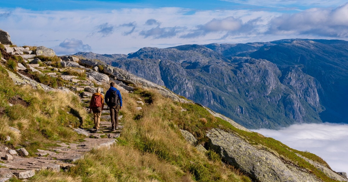 To personer går på en steinlagt fjellsti mot Kjerag, med bratte fjellformasjoner og skyer i dalen. Foto.