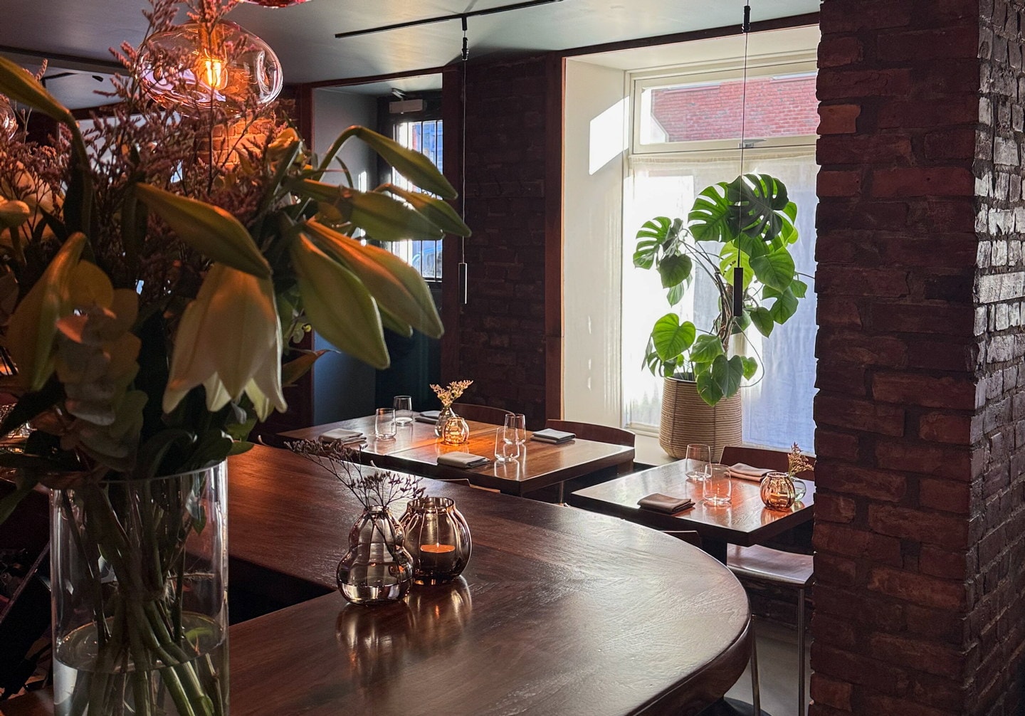 Restaurantinteriør med bardisk, blomster, hengelamper og dekkede bord ved et stort vindu.