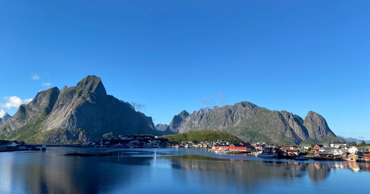 Bebyggelse ved sjøen omgitt av bratte fjell, med røde og hvite hus som speiler seg i vannet.