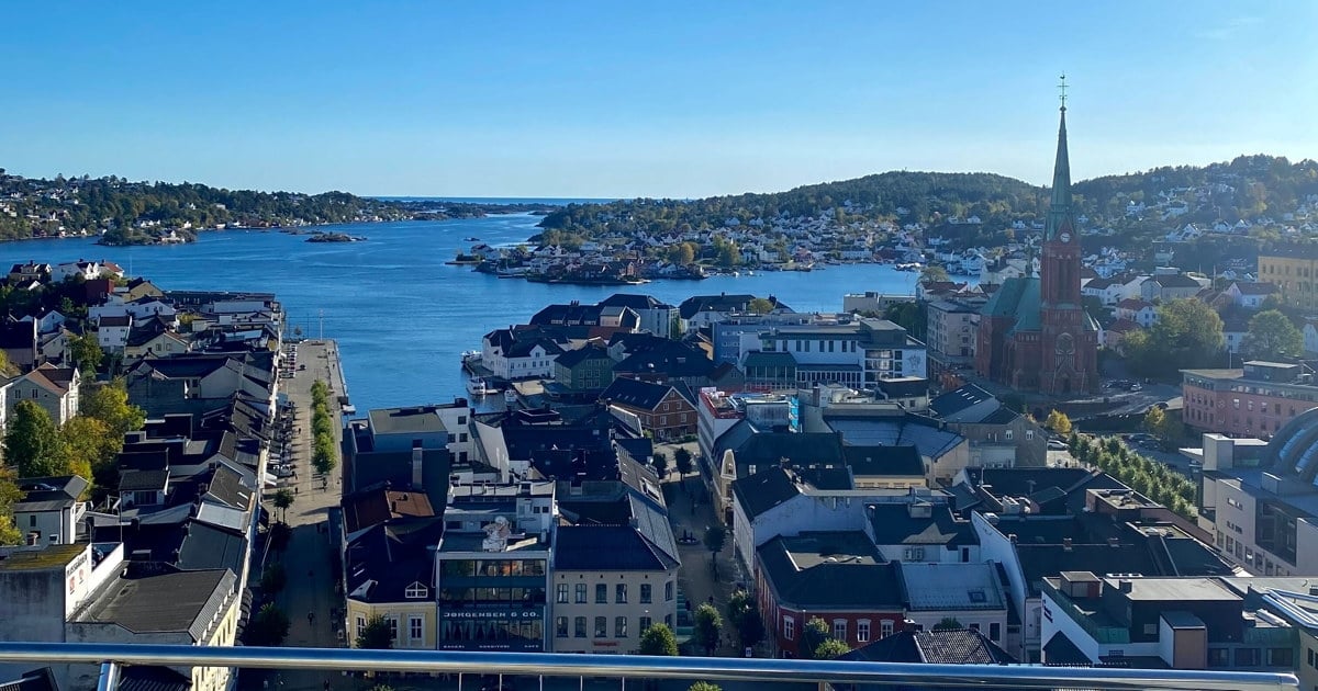 Utsikt over Arendal sentrum fra høyden, med Trefoldighetskirken, bebyggelse og sjøen under en klar, blå himmel.