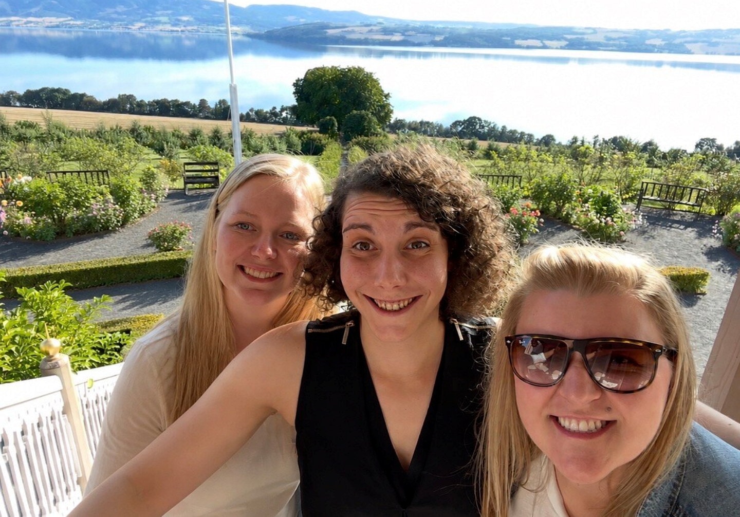 Tre personer tar en selfie utendørs med hage, vann og landskap i bakgrunnen.
