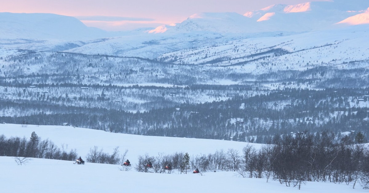 Snøscootere kjører gjennom et snødekt fjellandskap i vinterlys. Foto.