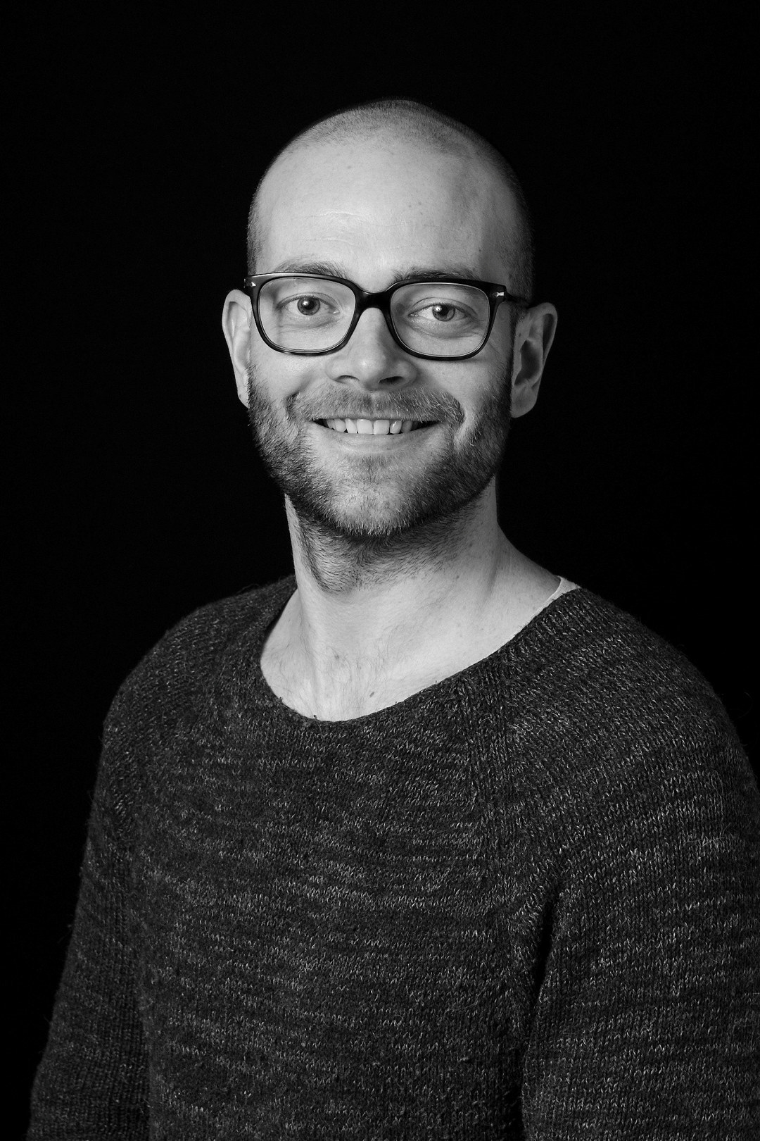 Et portrettbilde av en smilende mann med briller som ser mot kamera. Foto.