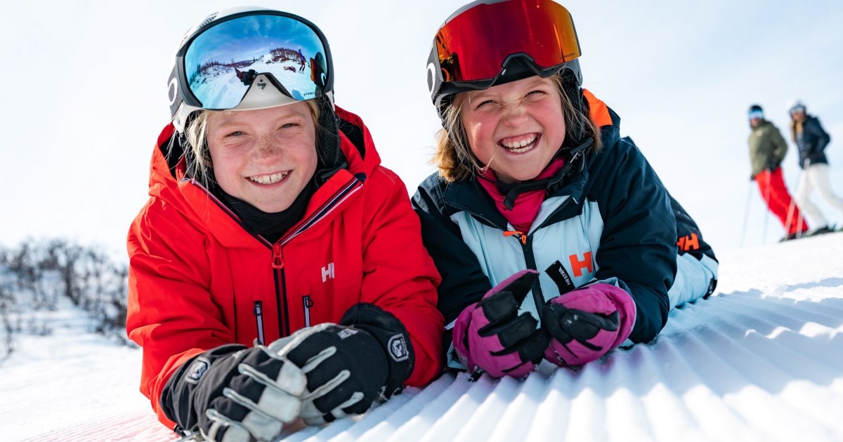 To barn med hjelm og slalåmbriller som ligger på magen i snøen
