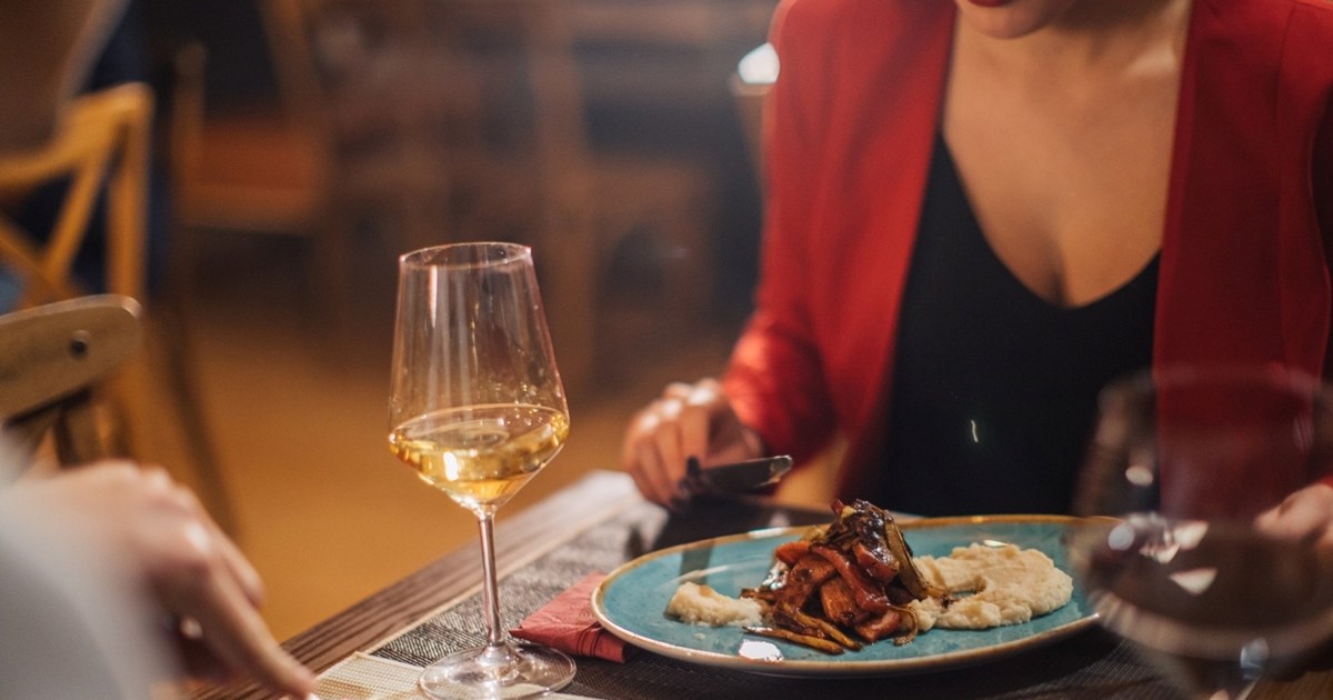 Foto av en kvinne som sitter ved et bord og spiser.  På bordet står et vinglass. Bildet viser kun utringingen hennes. Hodet er utenfor bildekanten.