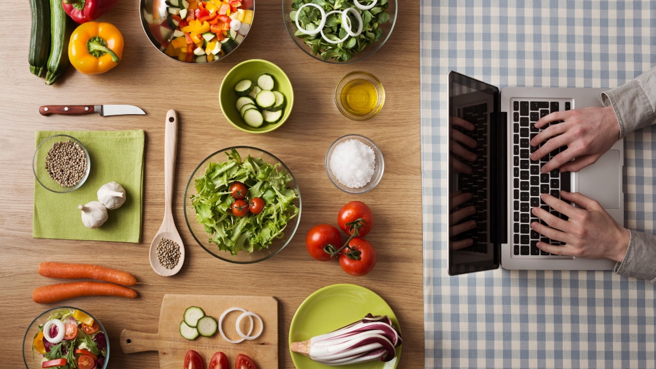 Fargerike frukt og grønnsaker i skåler på et bord. En datamaskin hvor en person sitter og skriver på enden av bordet.