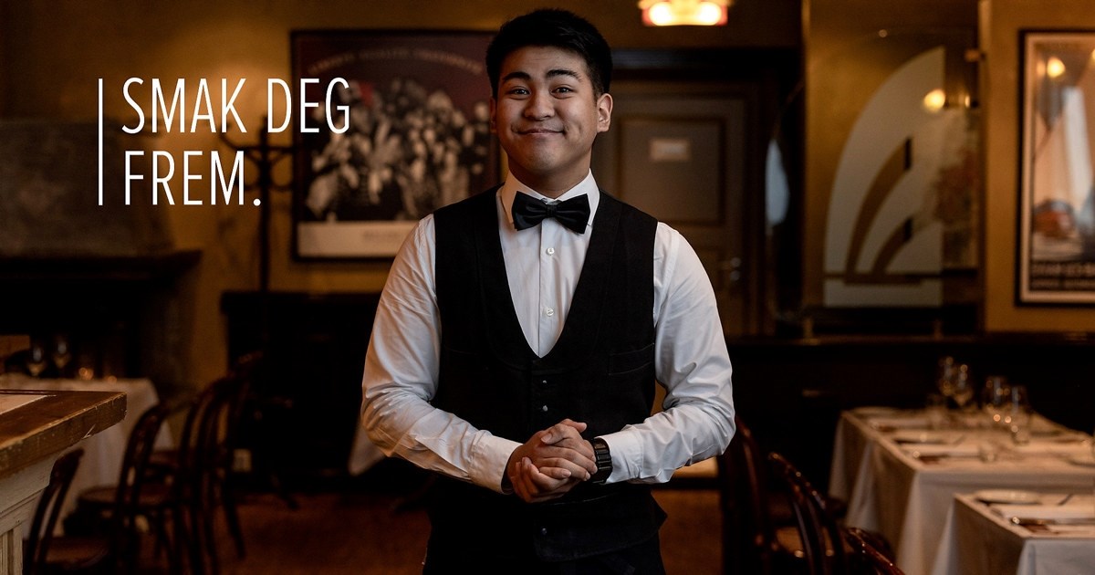 Ung, smilende mann i servitøruniform som står i en restaurant. Foto. Over bildet står teksten "Smak deg frem".