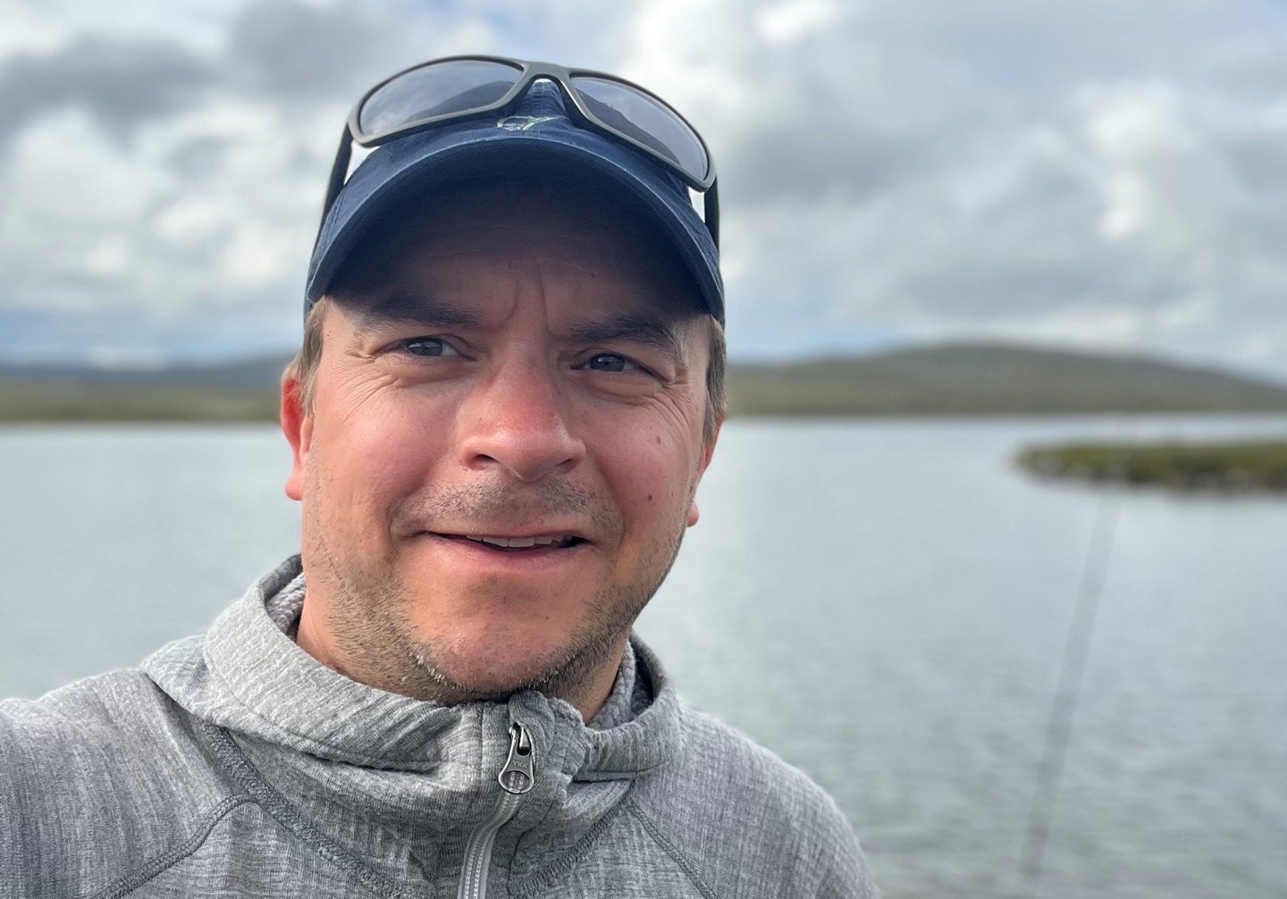 En mann med grå genser og solbriller oppå en blå caps. Foto.
