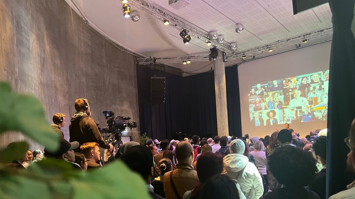 En mann som står på en scene foran veldig mange mennesker. To menn med videokamera står bakerst i lokalet. Foto