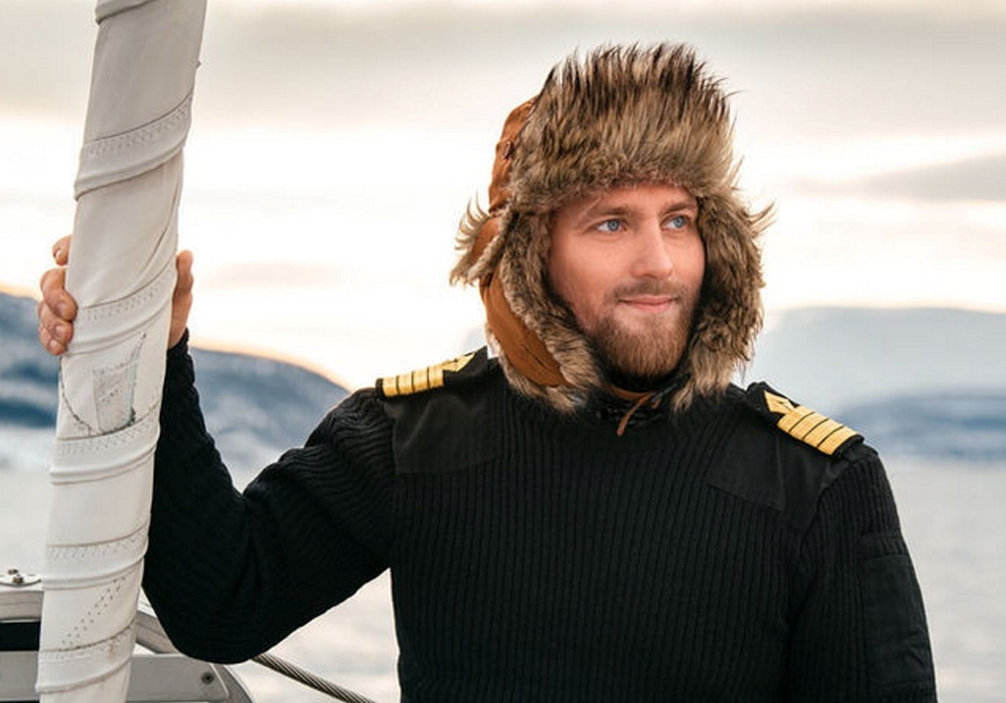 En mann med lue med pels og strikkegenser med kapteinsstriper på skuldrene, som står om bord i en båt. Foto