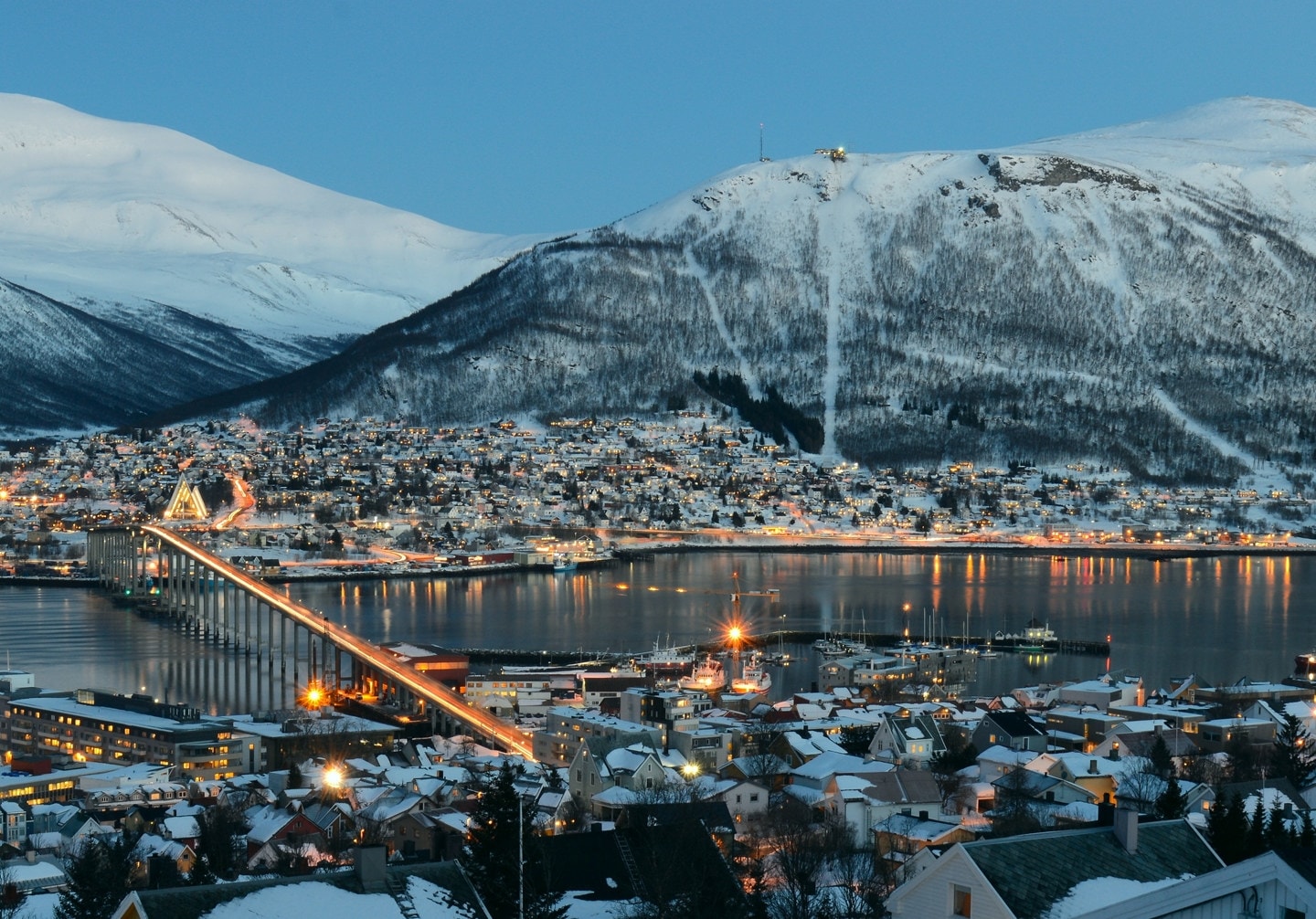 En by med mange hus ved vannet om kvelden, med snødekket fjell i bakgrunnen. Foto.