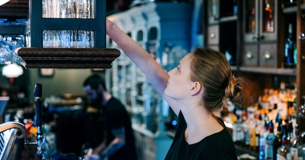 En kvinne som står bak en bardisk og strekker seg etter et glass i en hylle over baren. En annen person sees i bakgrunnen. Foto