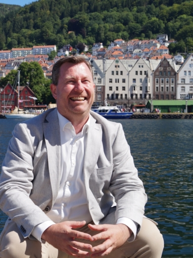 En smilende mann som sitter ved vannet og poserer mot kameraet, med hus på rekke i bakgrunnen. Foto.