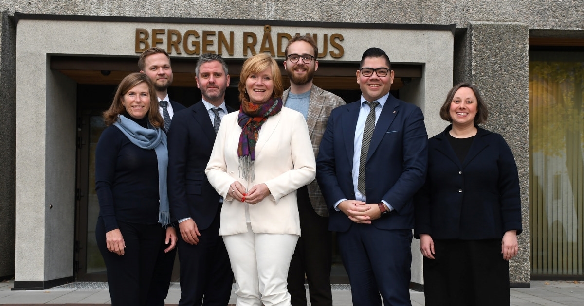 Sju mennesker som står på rekke foran en grå murbygning med skilting Bergen Rådhus bak seg. Foto