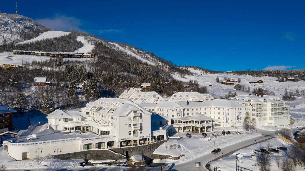 Bilde av et stort hvitt hotell, med skibakker, snø og blå himmel rundt. Foto.