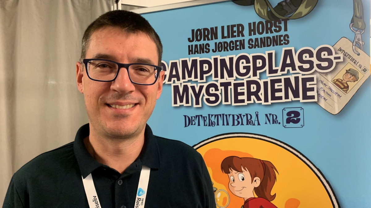 En mann med briller og konferansedeltaker-badge rundt halsen, som står foran en plakat som promoterer barneaktiviteten Campingplass-mysteriene. Foto.