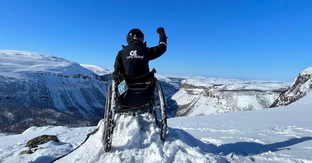 Naturbilde med en som sitter i rullestor på toppen av et fjell, med en hånd i været