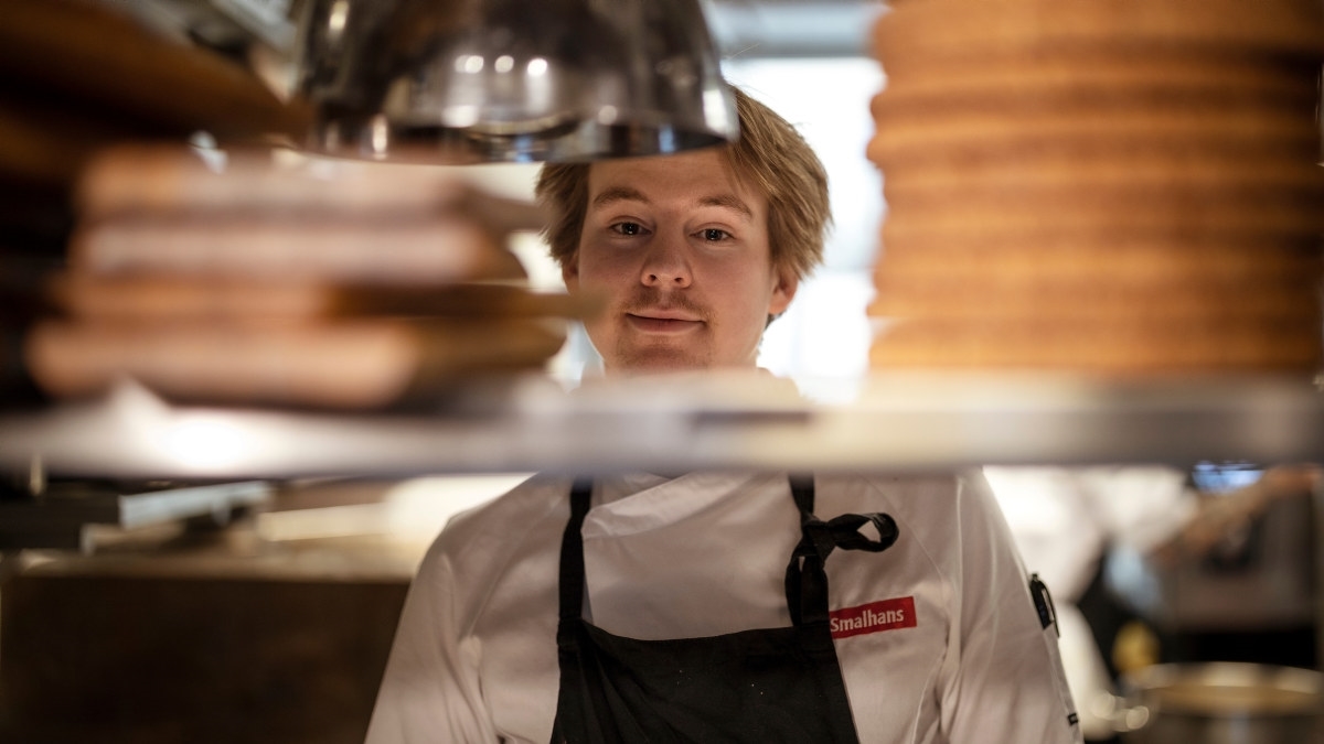 Ung kokk som titter mellom serveringshyller på et restaurantkjøkken. Foto