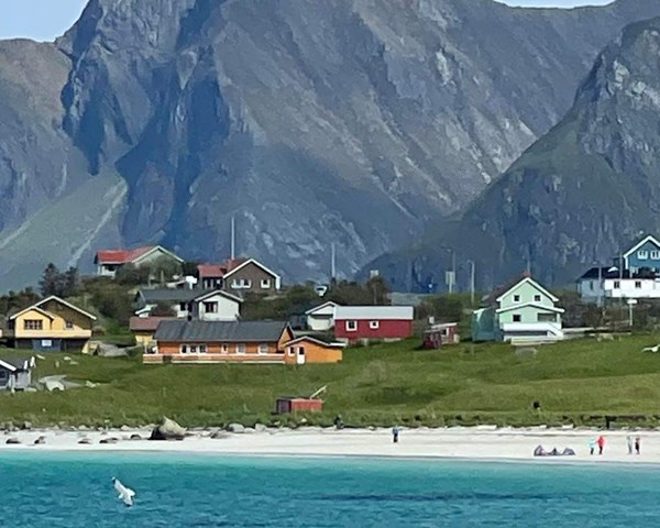 En strand med hav,  fjell og bebyggelse i ulike farger, i bakgrunnen. Foto