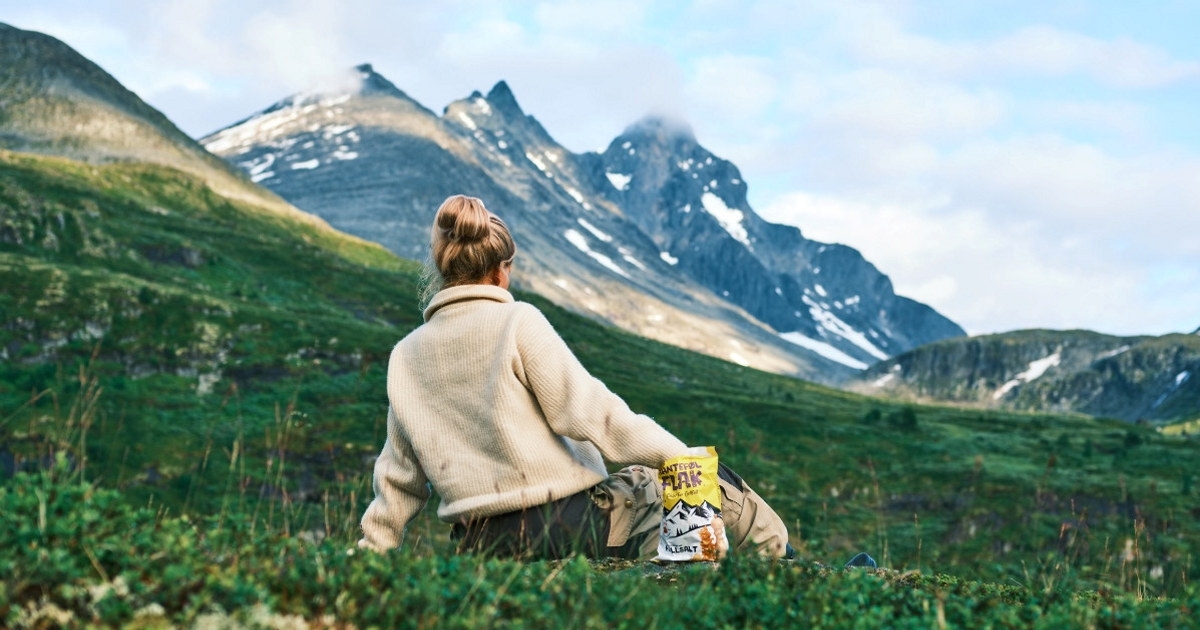 Dame som sitter i gress mellom fjell. Spiser fra en chipspose. Foto.
