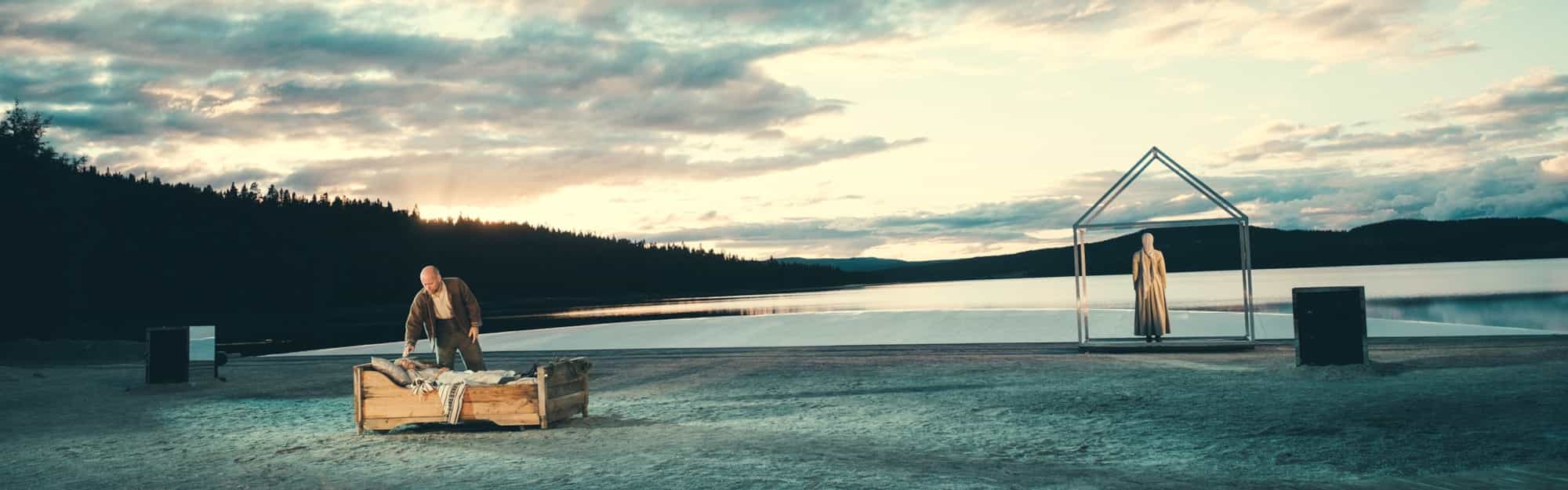 En strand der man ser en oppsetning av Peer Gynt. EN mann står over en seng med en kvinne i, mens en annen kvinne står til høyre i bildet, med ryggen mot. Foto